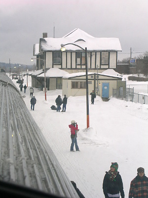 [Two-story station building and ground around it is completely snow-covered. People are scattered walking beside the train.]